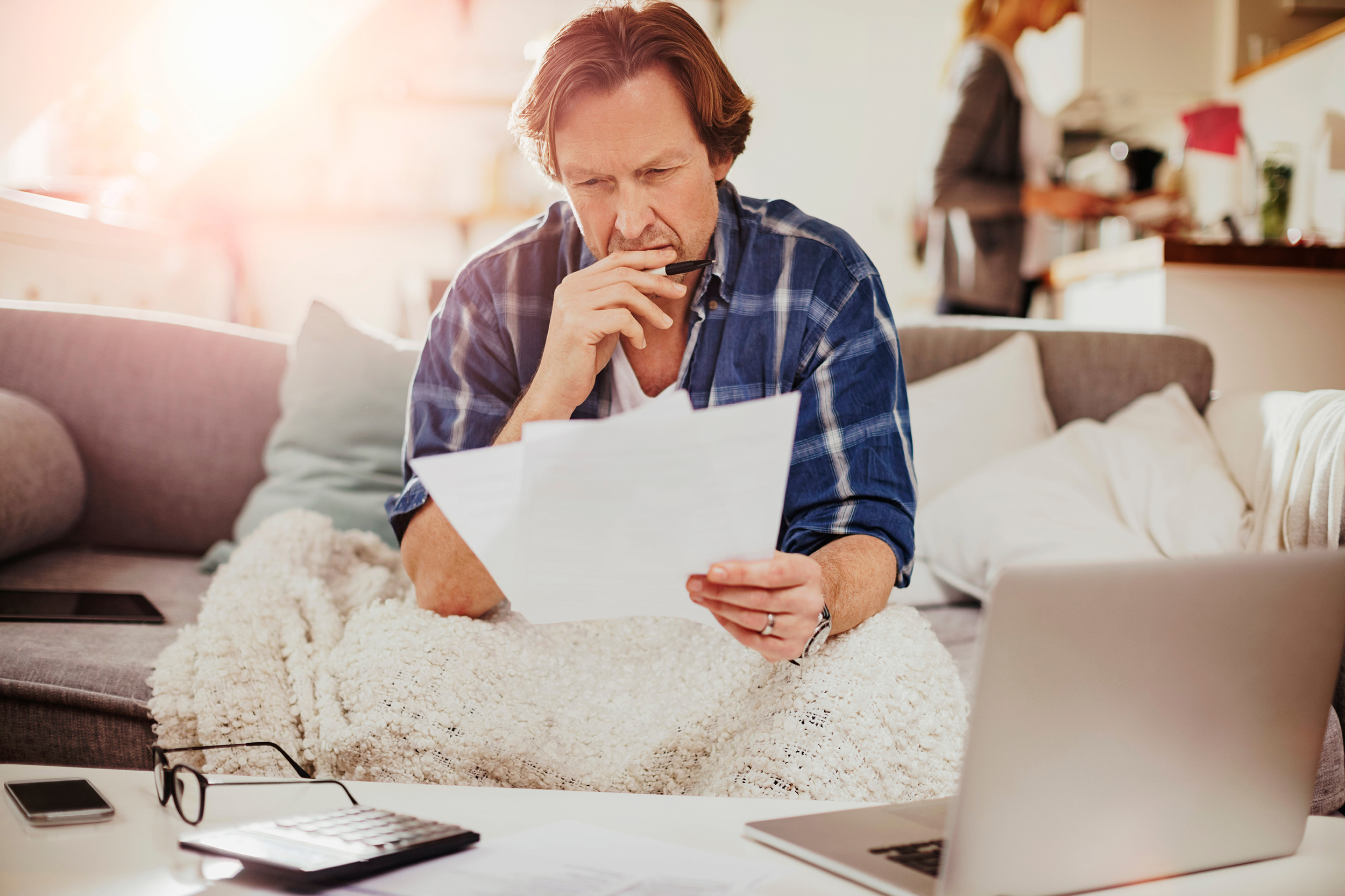 A man sits on a couch, blanket over his legs, studying papers in one hand as he puts his other hand, holding a pen, to his chin. West Virginians can get help determining if they have an industrial accident legal claim.