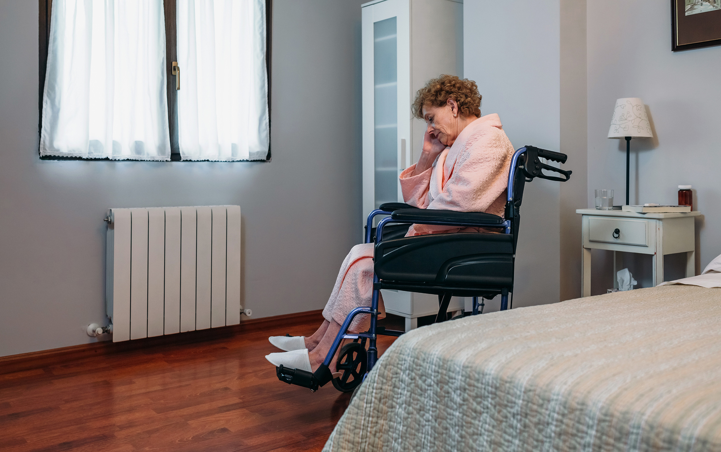 A woman sits in a wheelchair in her bedroom resting her head in her right hand. You can spot several possible signs of nursing home neglect and talk to Shaffer & Shaffer lawyers for help.